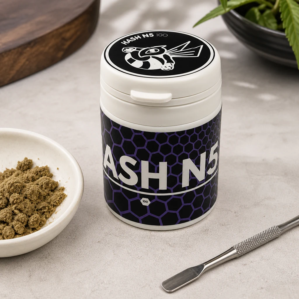Bote de Hash Dry N5 CBD Bee con tapa blanca y etiqueta de hexágonos morados, junto a un cuenco con material desmenuzado y una espátula.