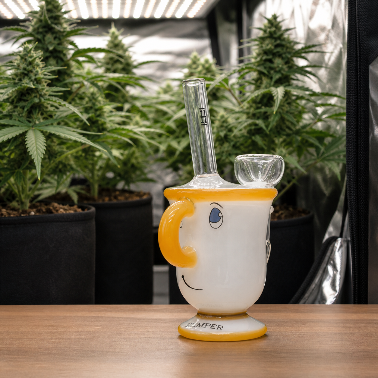 Bong Hemper en forma de taza blanca con asa y bordes amarillos, rostro sonriente, tubo y cazoleta transparentes, ante plantas de cannabis.