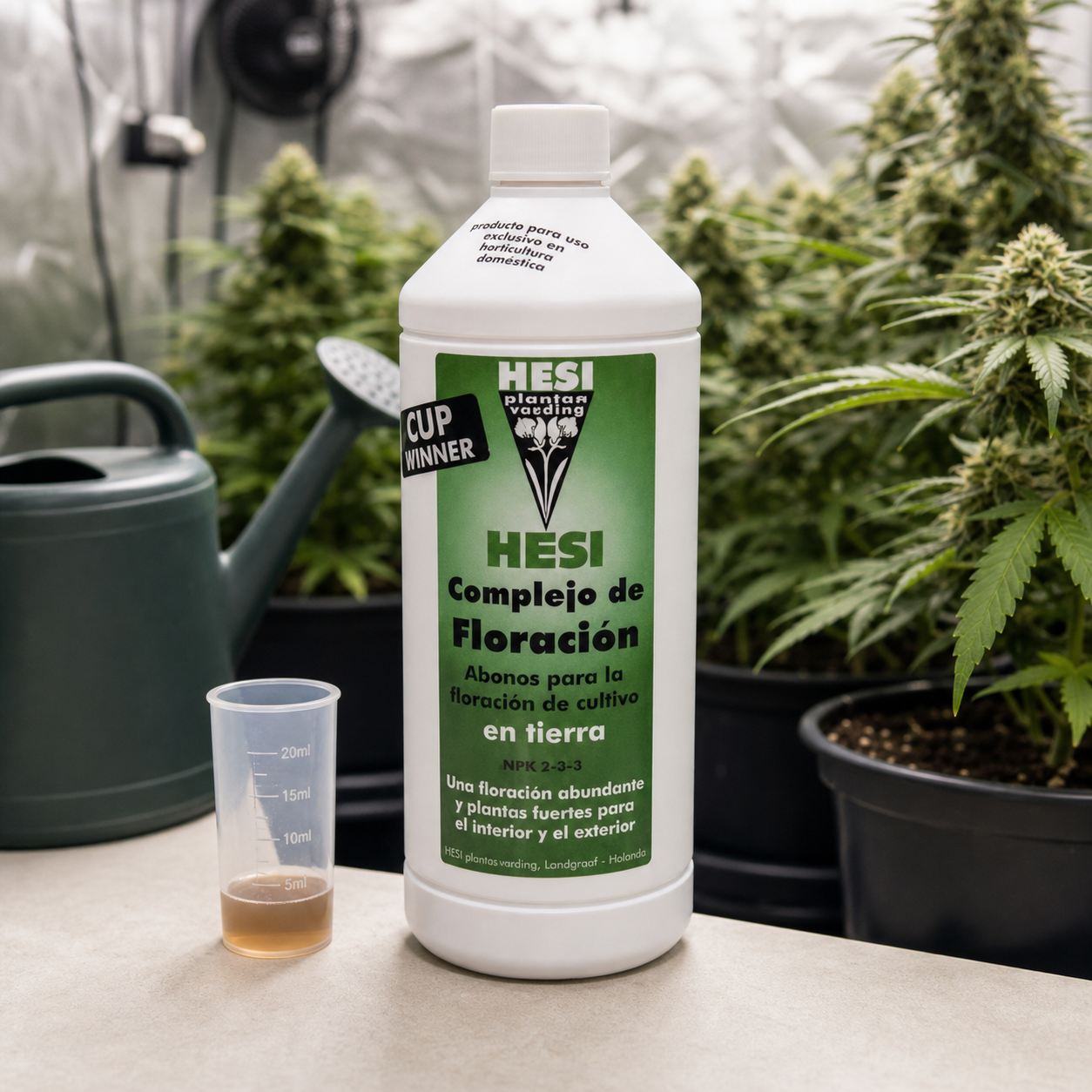 Botella de HESI Bloom Complex para suelo junto a un vaso dosificador con líquido y una regadera, ante plantas en floración.