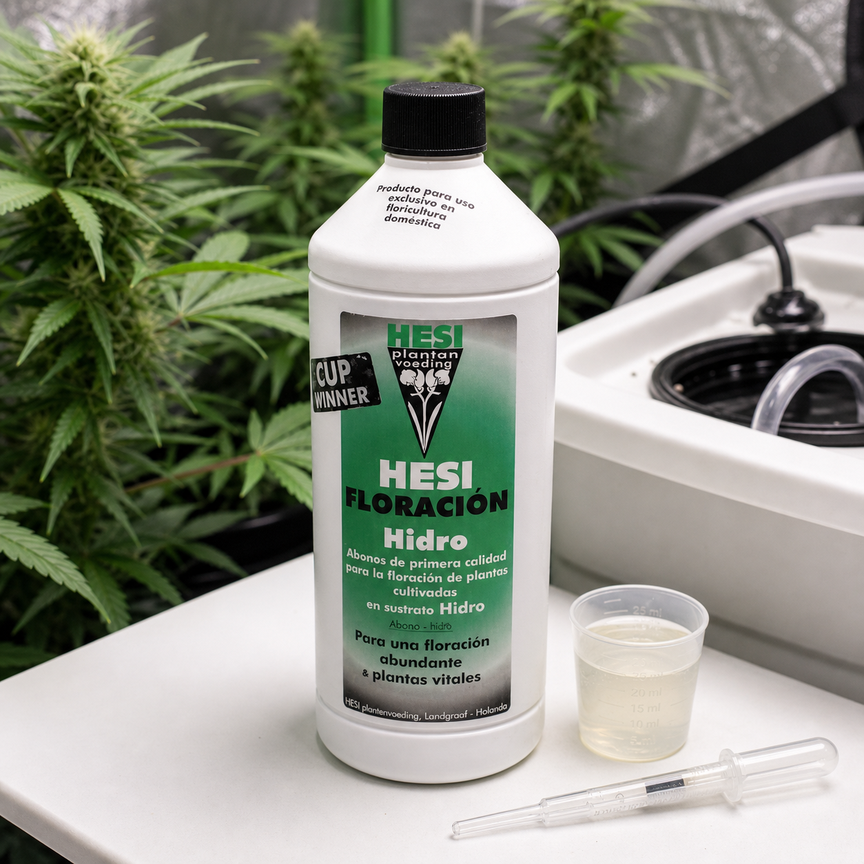 Botella de HESI Floración Hidro junto a un vaso dosificador y una pipeta, con plantas y un depósito con tubos al fondo.