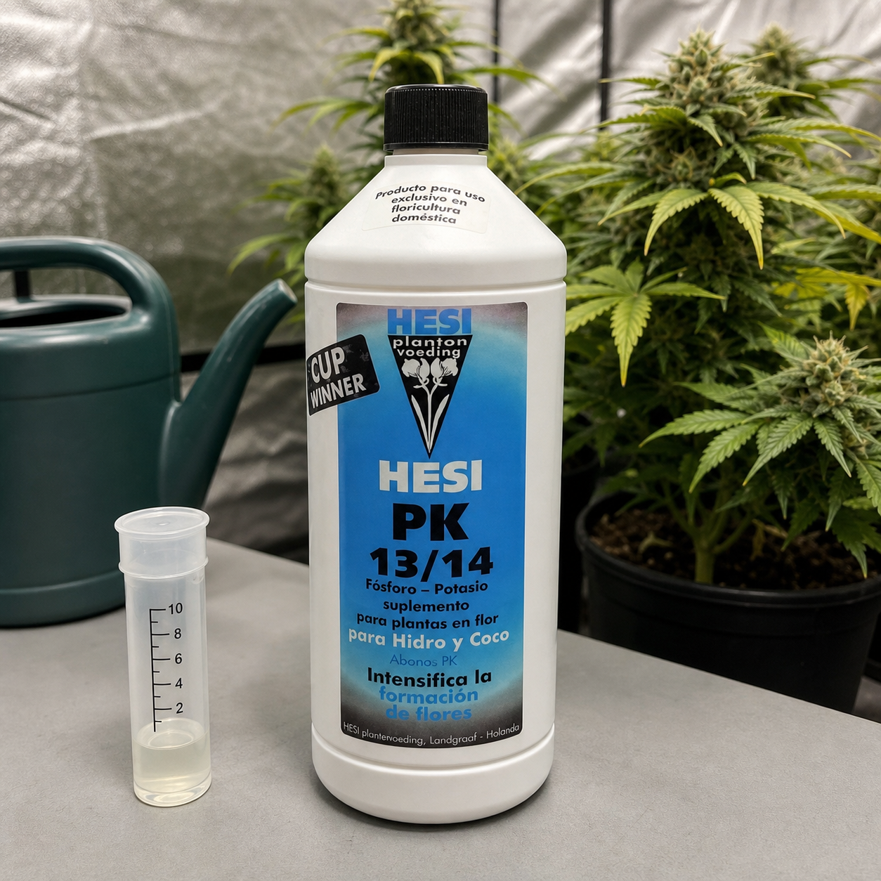 Botella HESI PK 13/14 con etiqueta azul y tapón negro junto a una probeta con líquido claro, con regadera y plantas en floración al fondo.