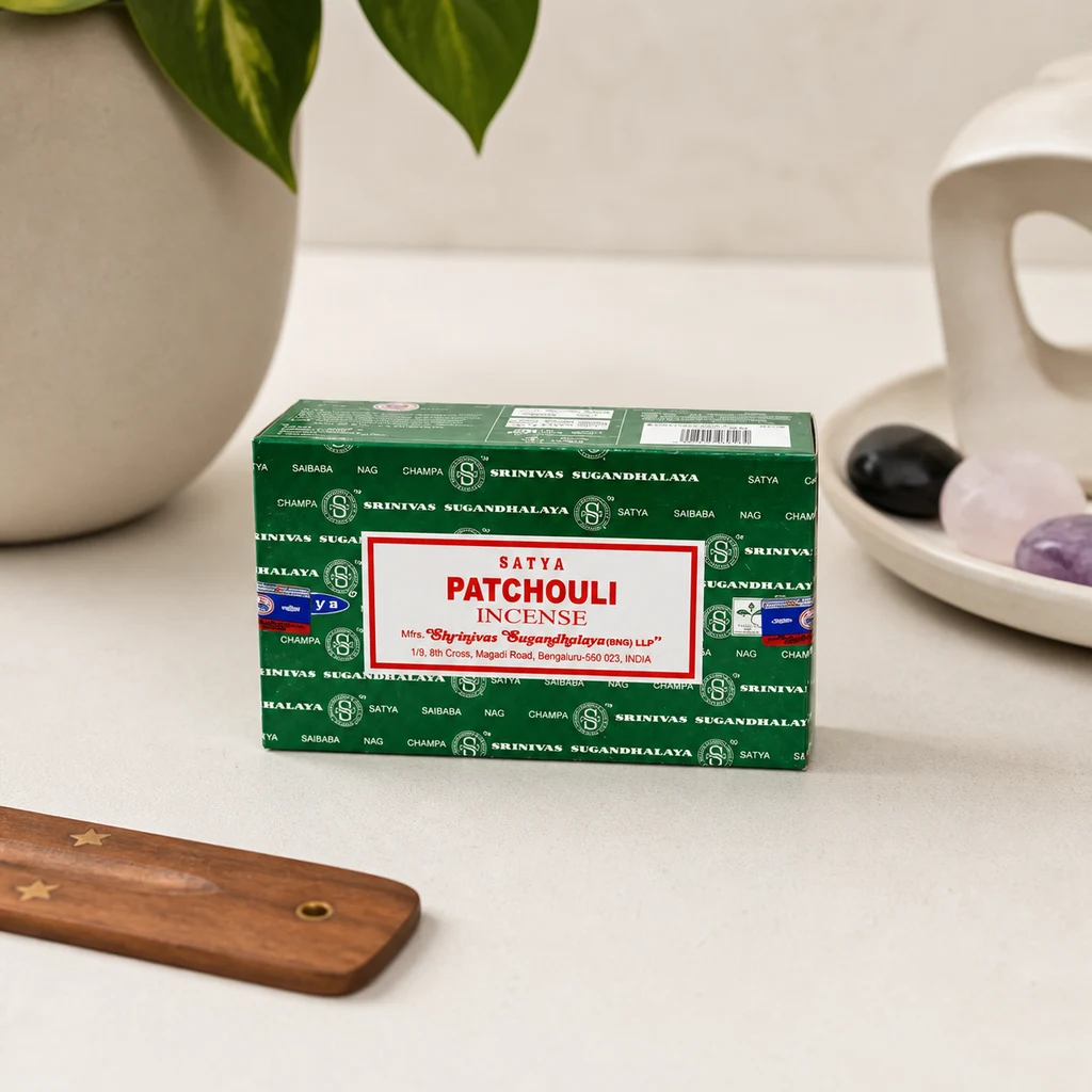 Caja verde de incienso Satya Patchouli sobre una superficie clara, junto a un soporte de madera, una maceta y una bandeja con piedras. Caja verde de incienso Satya Patchouli sobre una superficie clara, junto a un soporte de madera, una maceta y una bandeja con piedras.