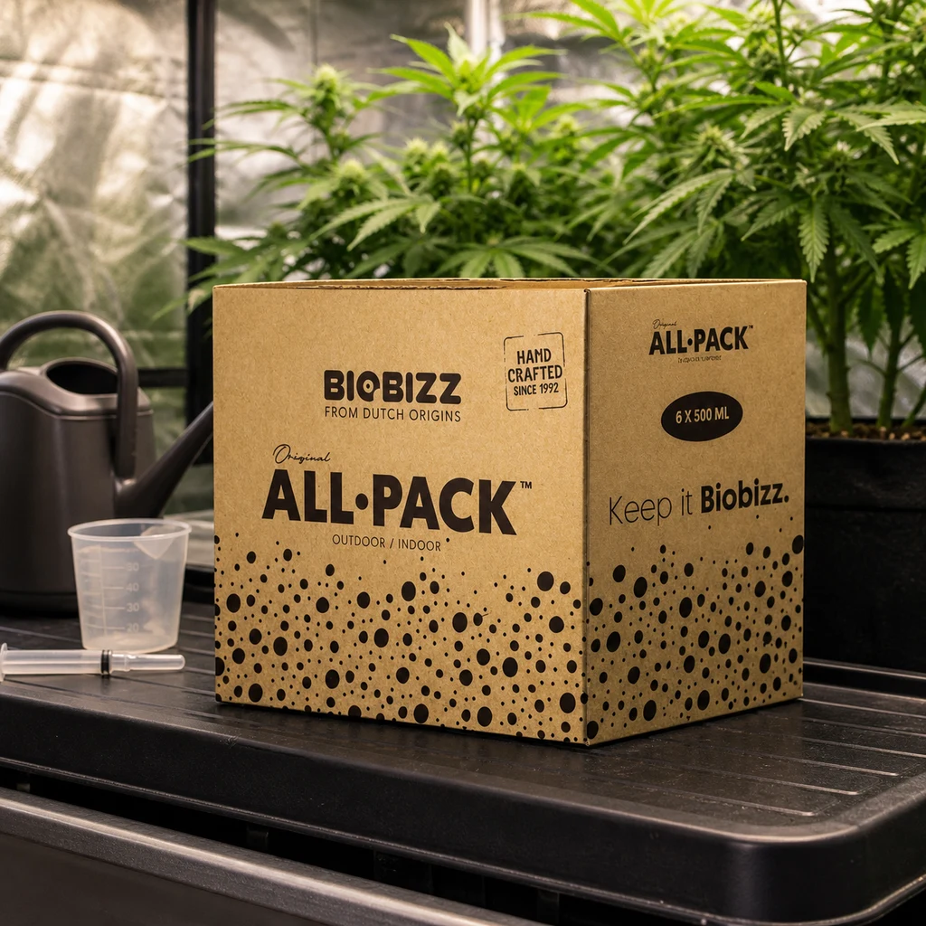 Caja BioBizz All-Pack rotulada «6 x 500 ml» sobre una bandeja, junto a una regadera y un vaso medidor, con plantas al fondo.