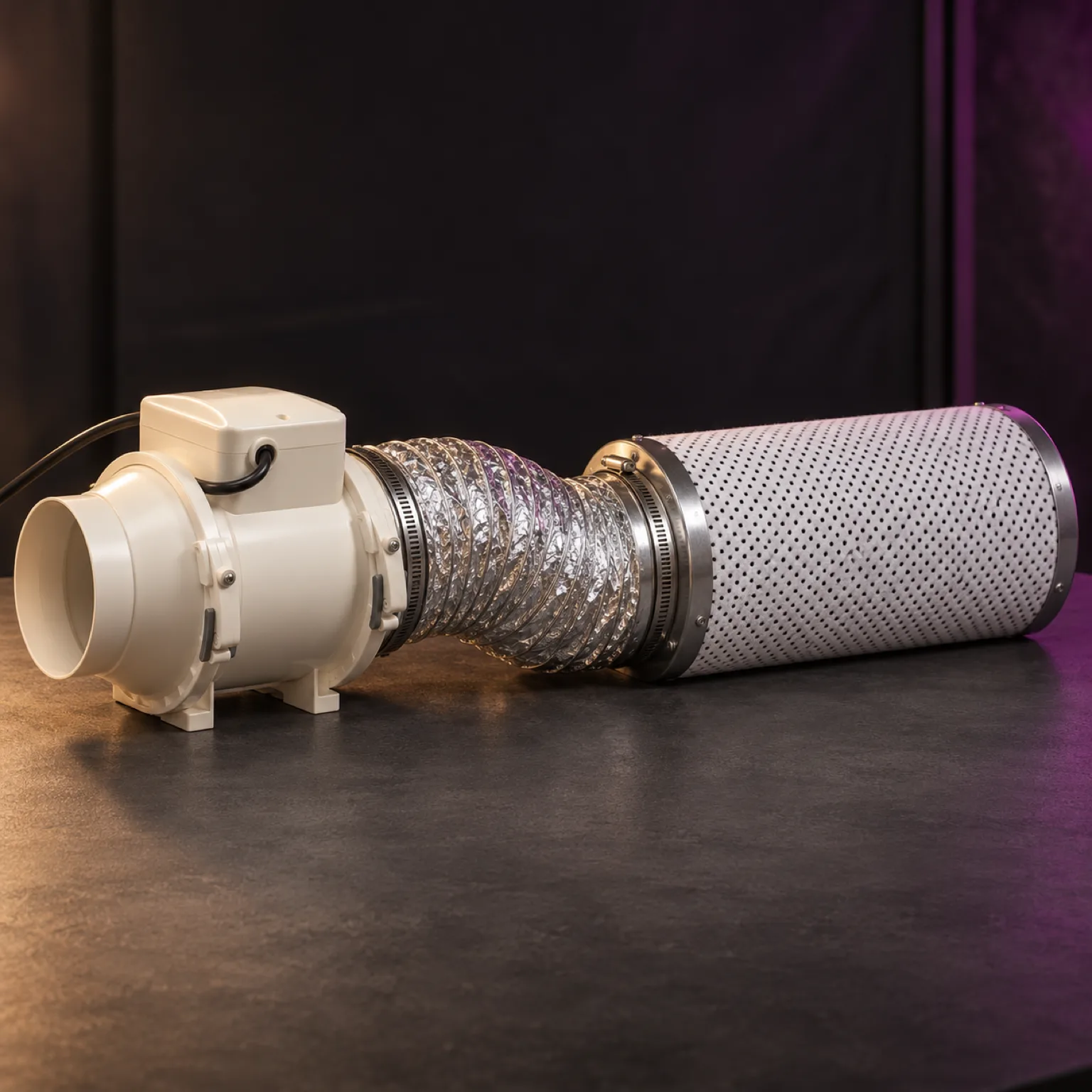 Kit de ventilación con extractor blanco conectado mediante un conducto flexible plateado a un filtro de carbón cilíndrico, sobre una mesa oscura.