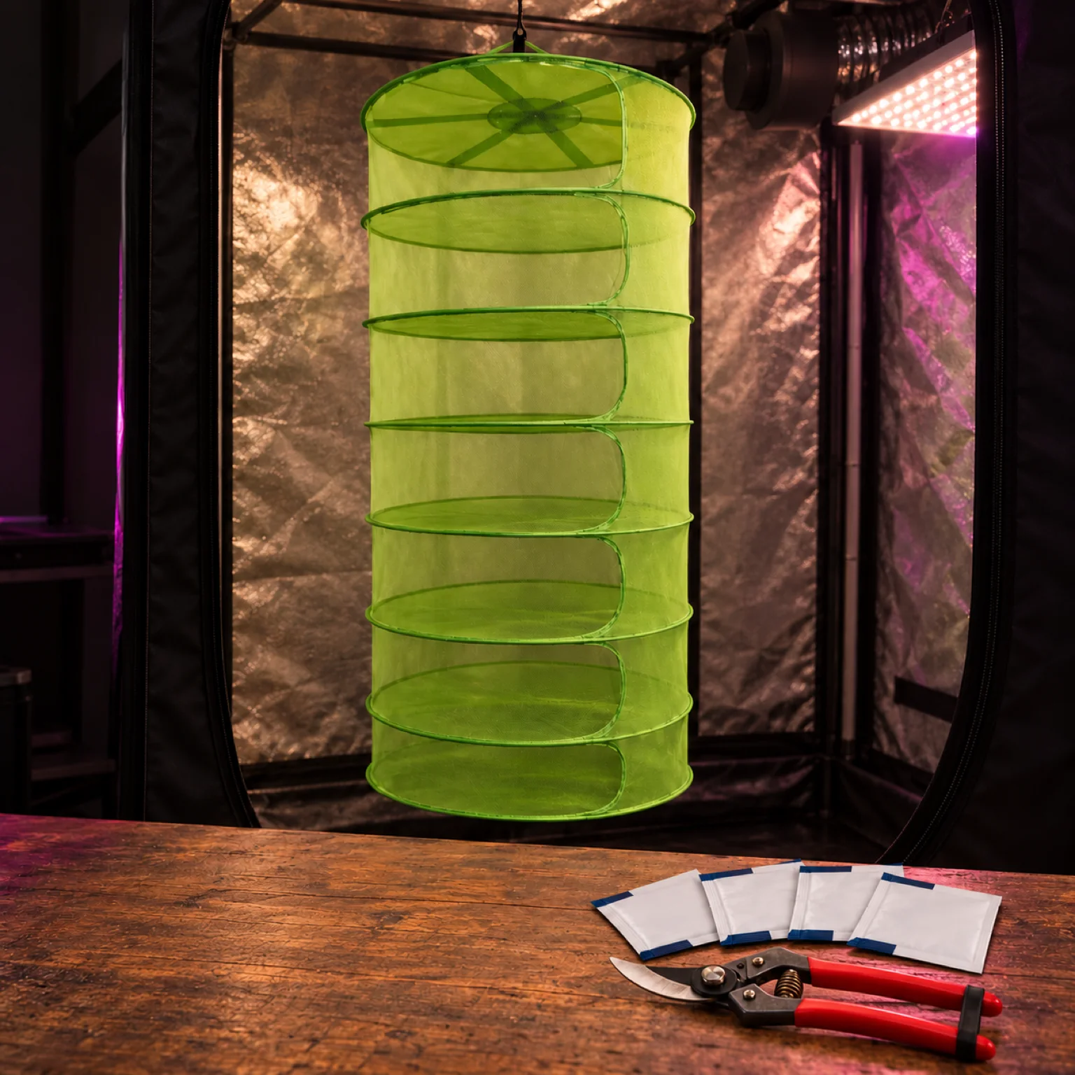 Secadero colgante verde de varios niveles en un armario de cultivo, con tijeras de poda y varios sobres sobre una mesa.