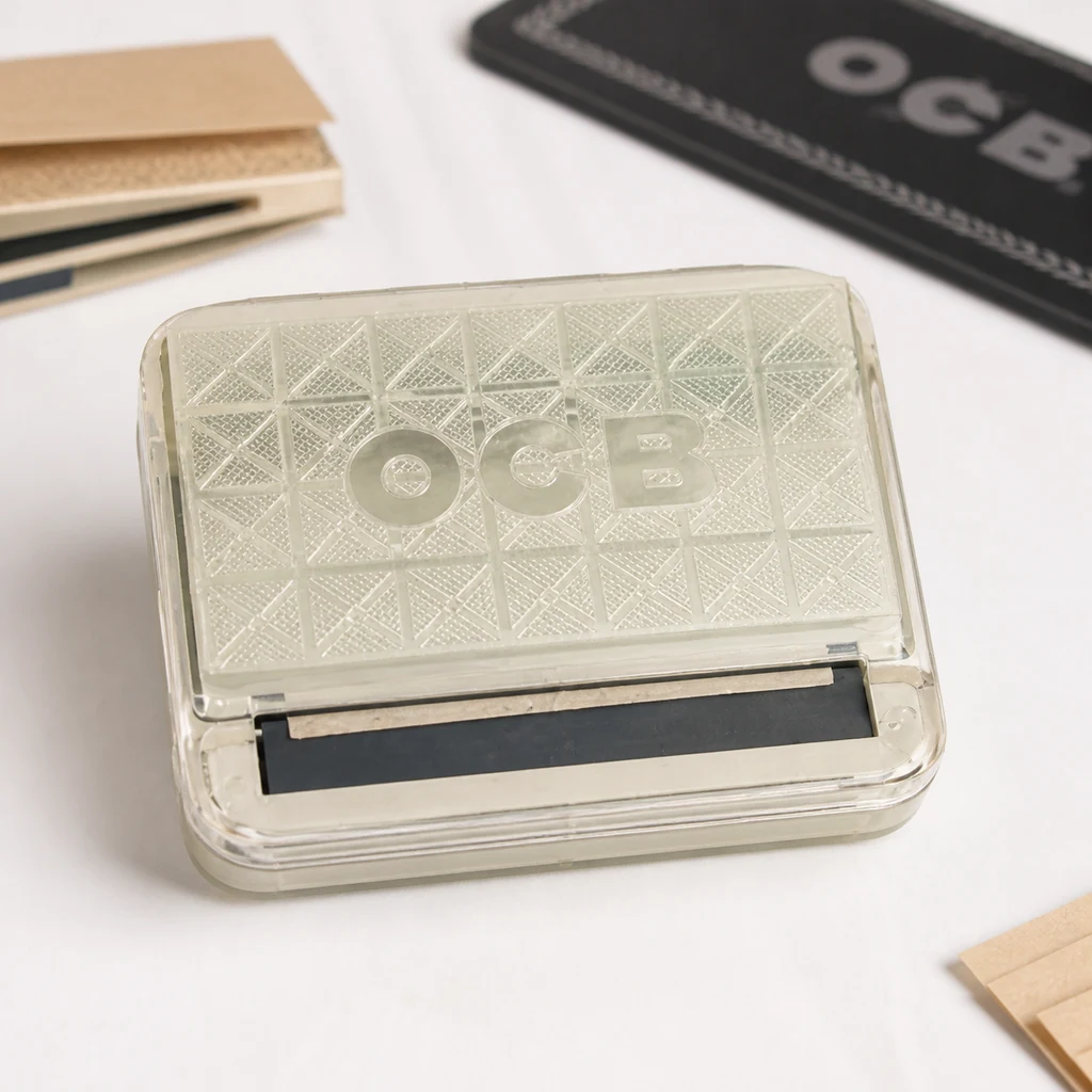 Caja de liar OCB cerrada, con tapa translúcida de relieve geométrico y rodillo negro frontal, sobre una mesa con papelillos. Caja de liar OCB cerrada, con tapa translúcida de relieve geométrico y rodillo negro frontal, sobre una mesa con papelillos.