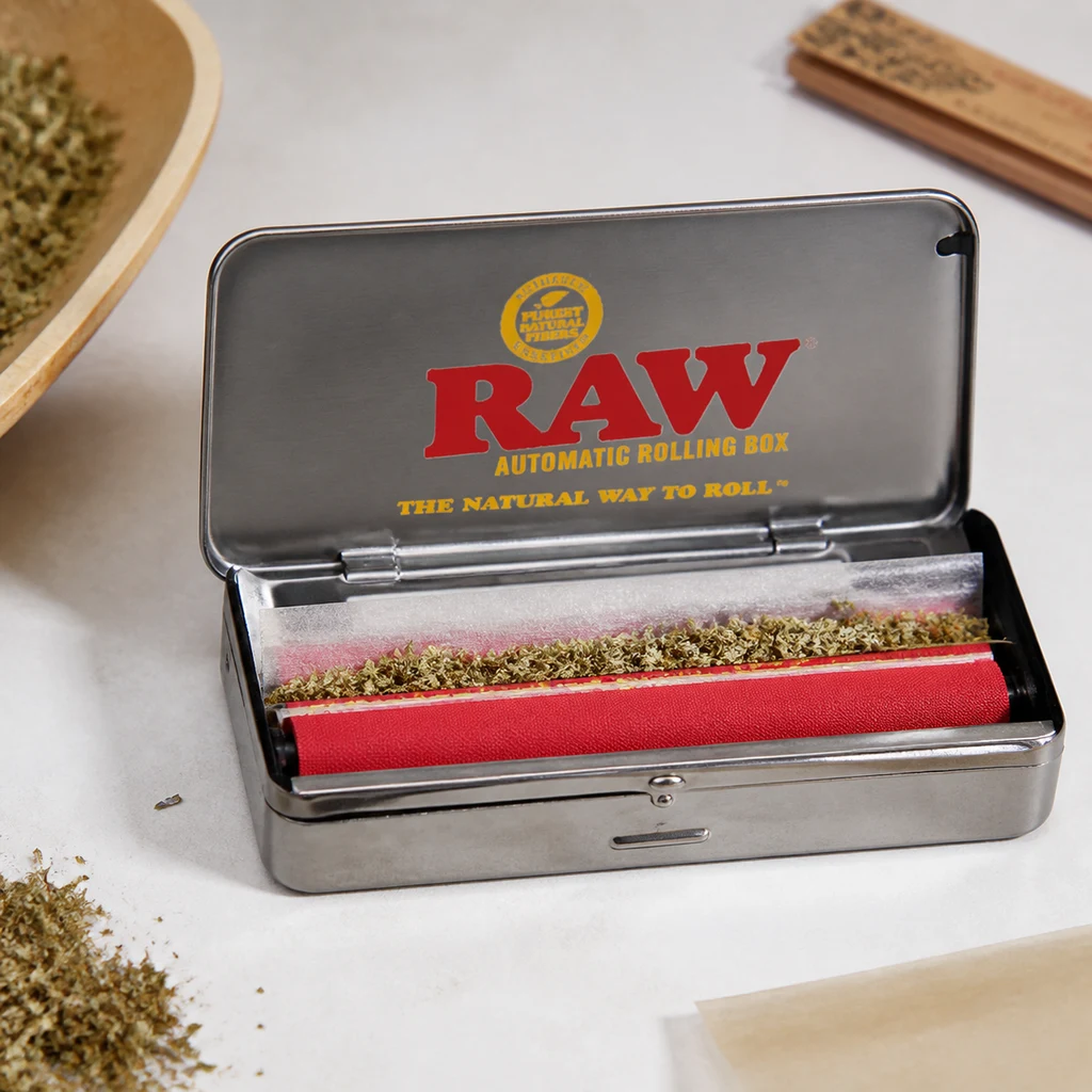 Máquina de liar RAW abierta, con rodillo rojo y papel con material vegetal desmenuzado; el logotipo aparece en el interior de la tapa. Máquina de liar RAW abierta, con rodillo rojo y papel con material vegetal desmenuzado; el logotipo aparece en el interior de la tapa.