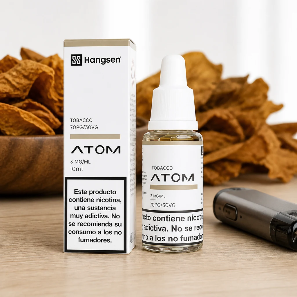 Frasco y caja de Hangsen Atom Tobacco de 10 ml y 3 mg/ml, sobre una mesa con hojas secas y un dispositivo de vapeo al lado.