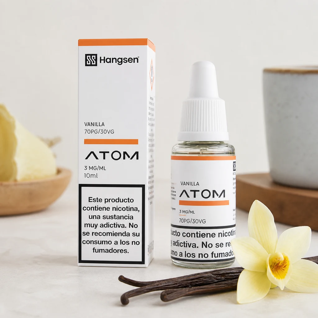 Frasco y caja Hangsen Atom Vanilla de 10 ml, etiquetados 3 mg/ml y 70PG/30VG, junto a vainas y una flor de vainilla, con una taza al fondo.