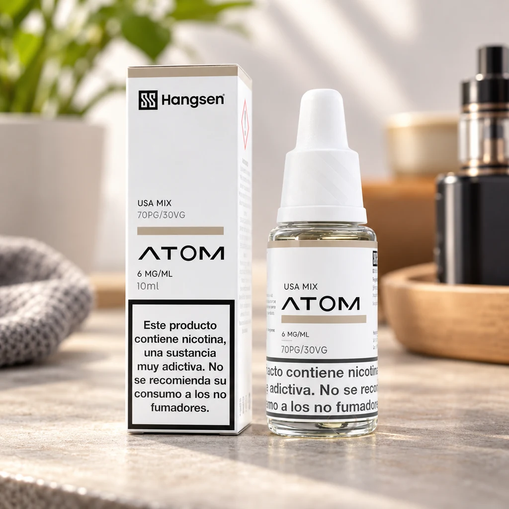 Frasco y caja de Hangsen Atom USA Mix sobre una mesa, con etiqueta de 6 mg/ml y advertencia de nicotina; vapeador desenfocado al fondo.