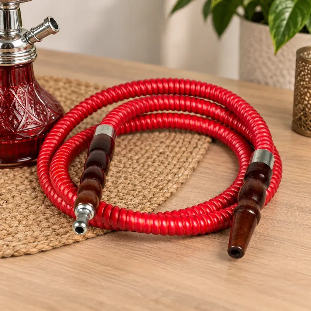 Manguera roja de cachimba enrollada, con extremos marrones y conexiones plateadas, sobre una mesa junto a una cachimba roja parcialmente visible.