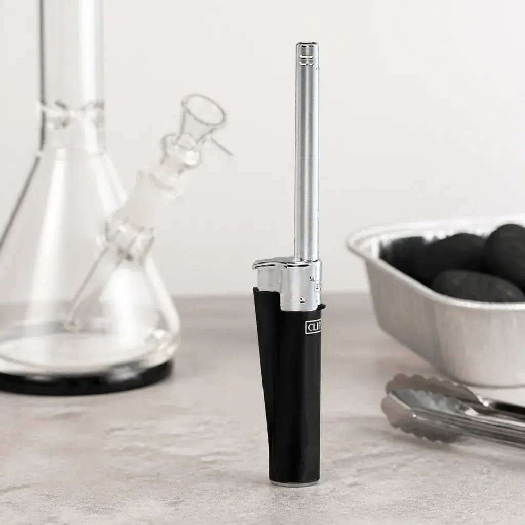 Encendedor Clipper de cuerpo negro y tubo plateado largo, en posición vertical junto a un bong, una bandeja con carbón y unas pinzas. Encendedor Clipper de cuerpo negro y tubo plateado largo, en posición vertical junto a un bong, una bandeja con carbón y unas pinzas.