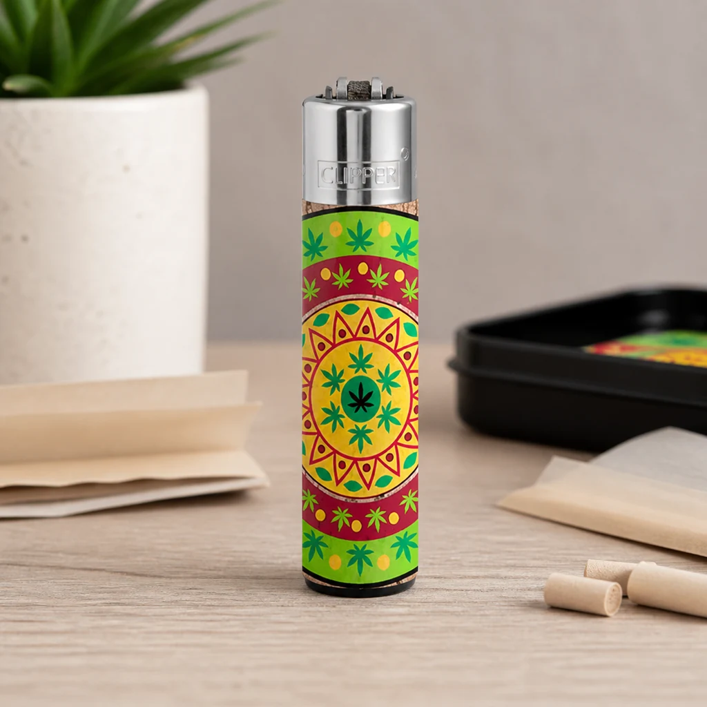 Encendedor Clipper con mandala y hojas verdes sobre fondo amarillo y rojo, de pie en una mesa junto a papeles y una bandeja.