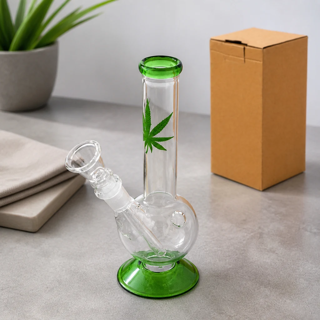 Mini bong de vidrio transparente con base y borde verdes, hoja de cannabis decorativa y cazoleta lateral sobre una mesa gris.
