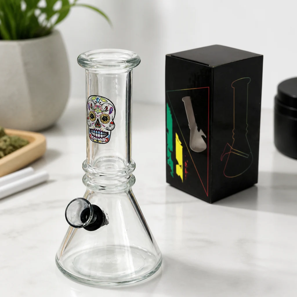 Bong transparente con base cónica, cazoleta lateral y calavera mexicana de colores, sobre una mesa junto a una caja negra.