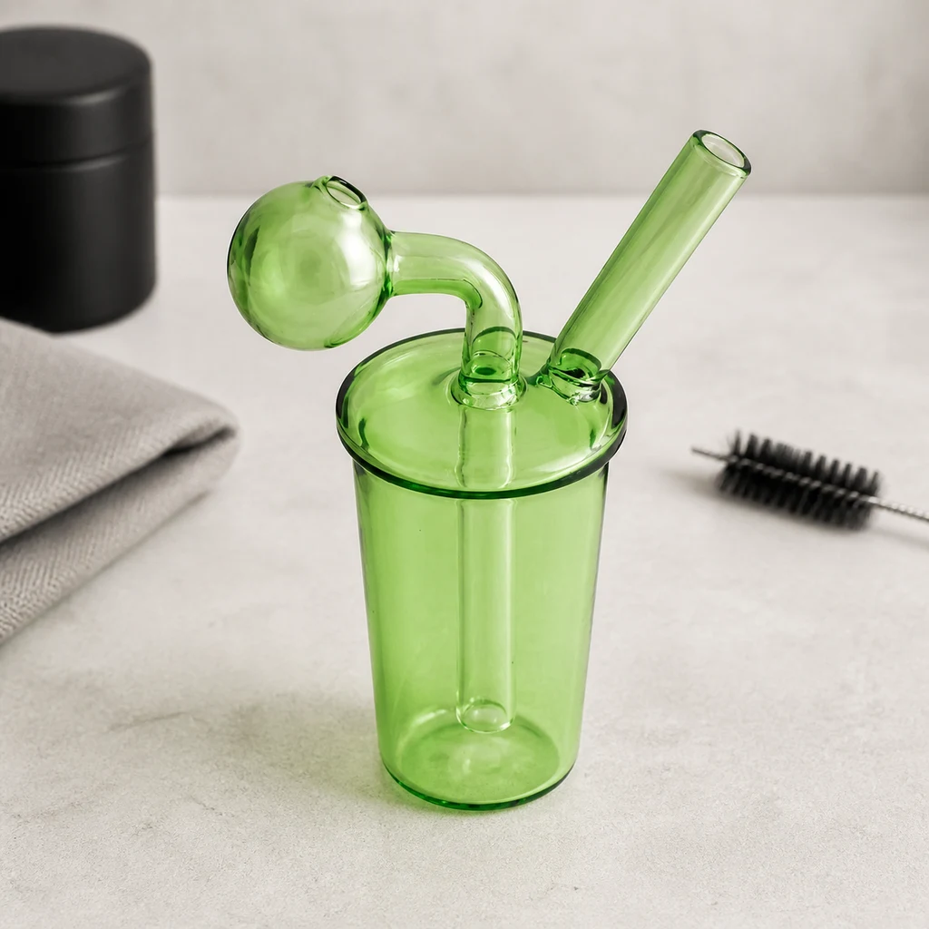 Mini bong verde translúcido con depósito cilíndrico, cazoleta esférica y boquilla inclinada, junto a un paño y un cepillo.