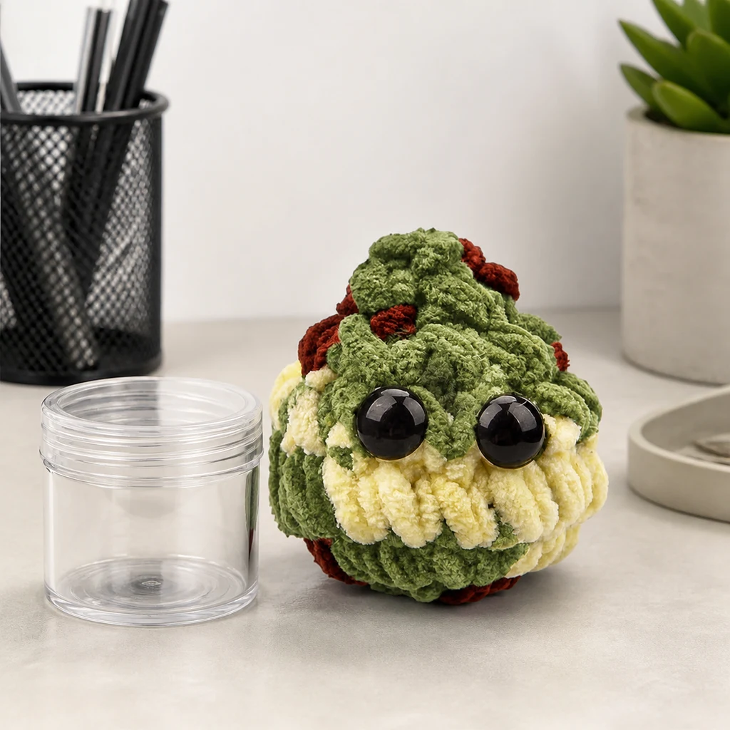 Mini Plush Bud verde con detalles crema y rojizos y ojos negros brillantes, junto a un frasco transparente abierto sobre una mesa. Mini Plush Bud verde con detalles crema y rojizos y ojos negros brillantes, junto a un frasco transparente abierto sobre una mesa.
