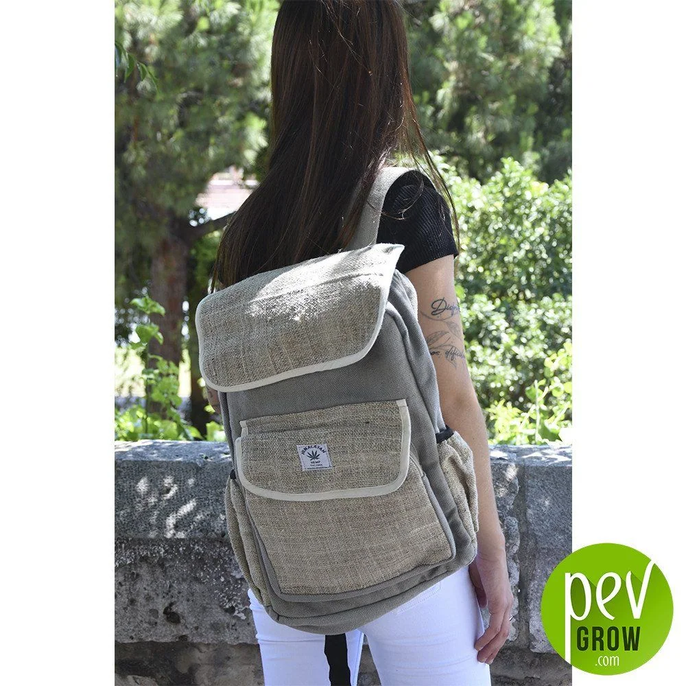Persona de espaldas con mochila gris y beige, con solapa amplia, bolsillo frontal y bolsillos laterales, en un entorno arbolado.