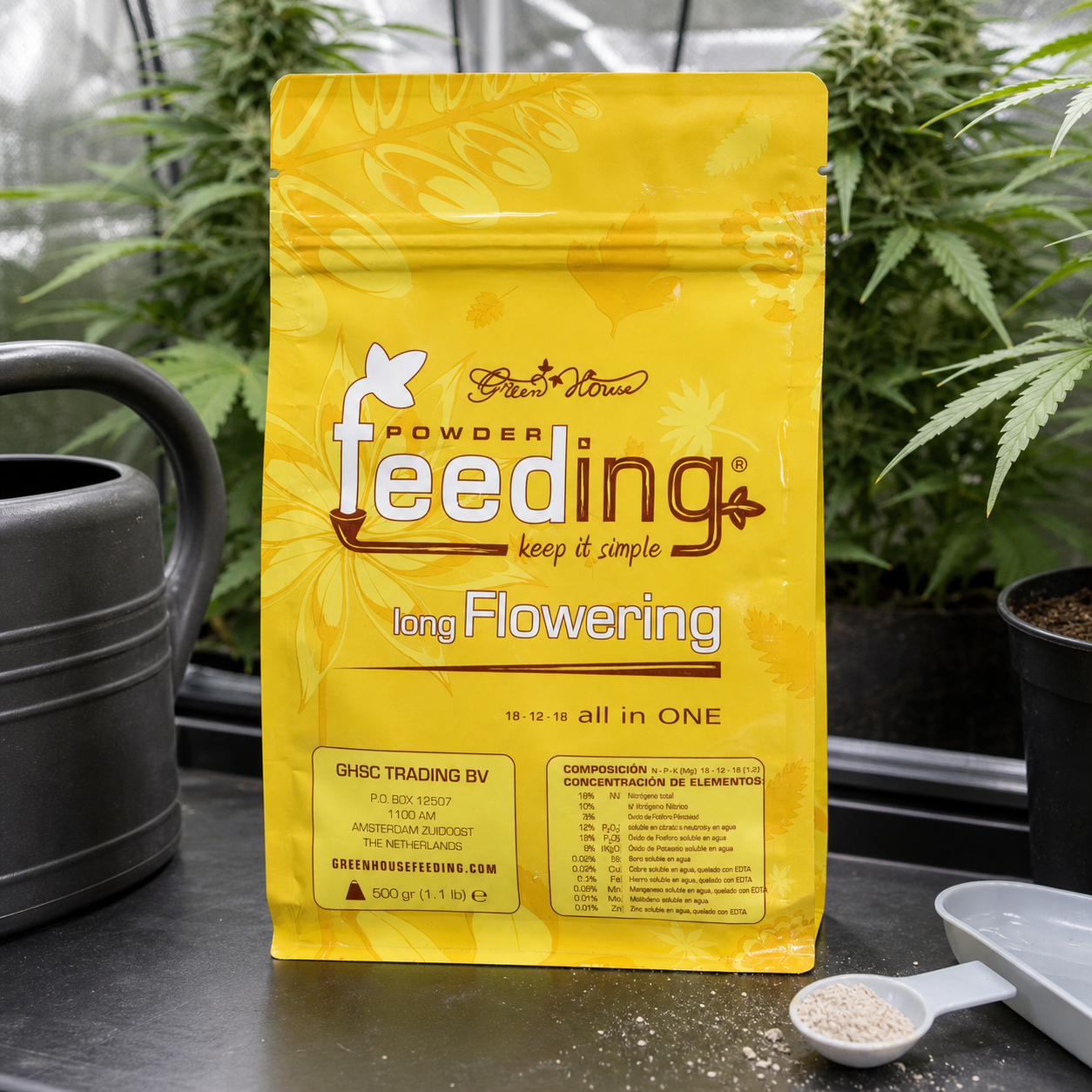 Bolsa amarilla de Green House Feeding Long Flowering junto a una cuchara con granulado, una regadera y plantas al fondo.