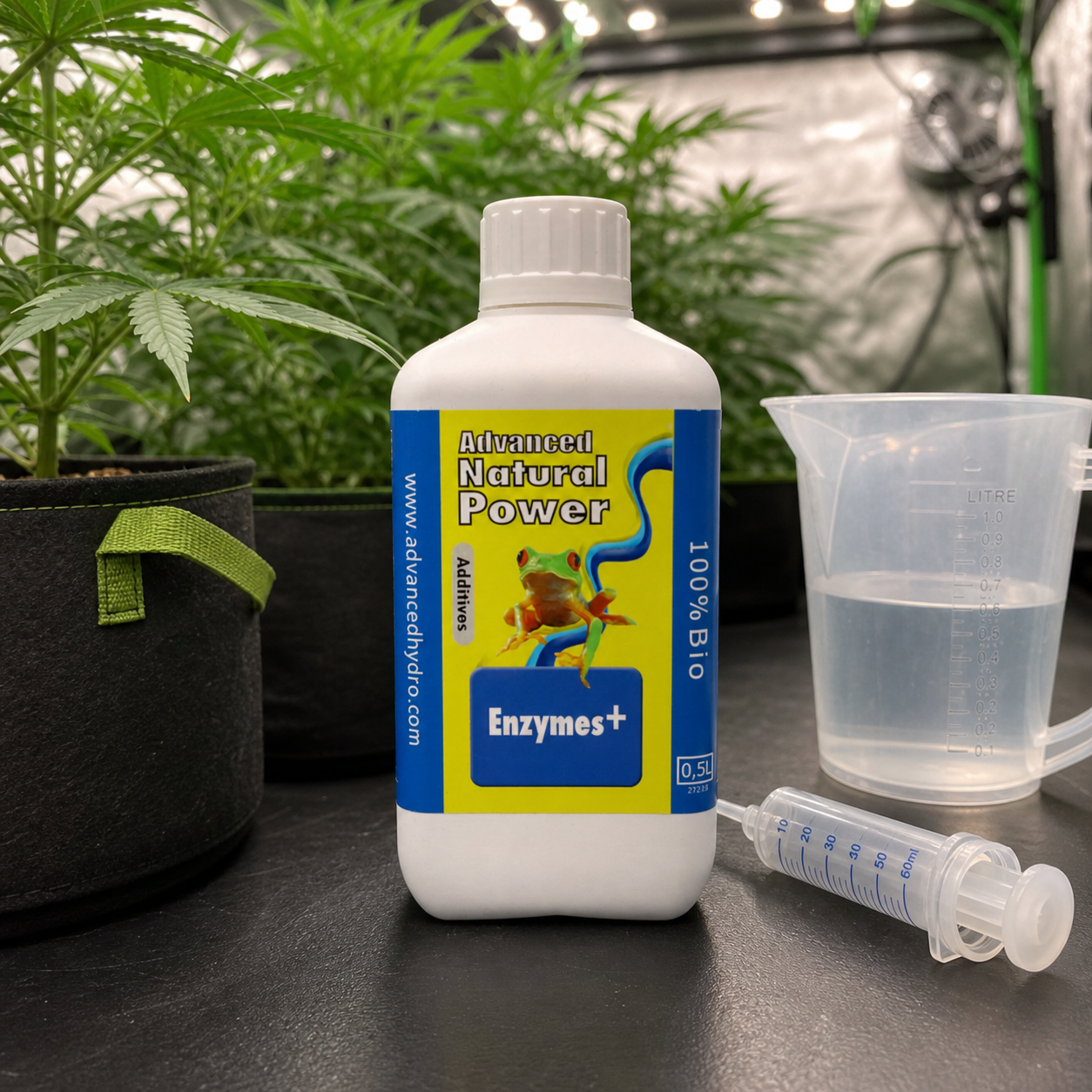 Botella de Natural Power Enzymes+ con etiqueta azul y amarilla, junto a una jarra con líquido y una jeringa, ante plantas de cannabis.