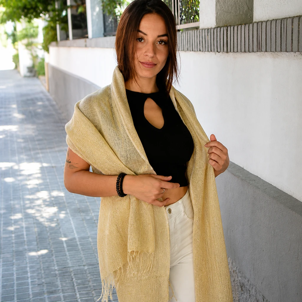 Mujer con un fular beige de tejido abierto y flecos sobre los hombros, camiseta negra y pantalón blanco, en una acera.