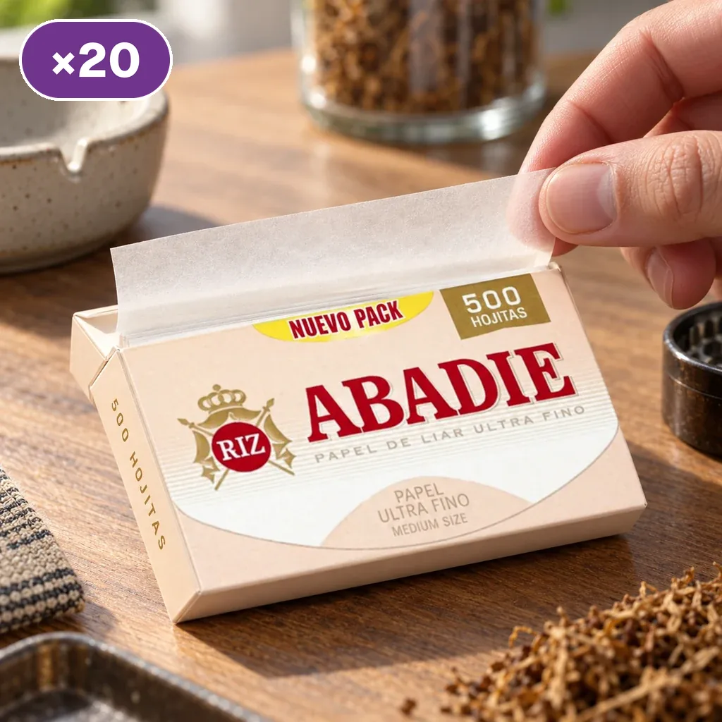 Una mano extrae una hoja de un bloque Abadie de 500 hojitas sobre una mesa, junto a un cenicero y un grinder; distintivo ×20.