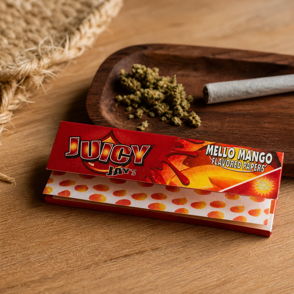 Librillo abierto de papeles Juicy Jay's Mello Mango sobre una mesa de madera, con una bandeja de hierba desmenuzada y un cigarrillo liado detrás.