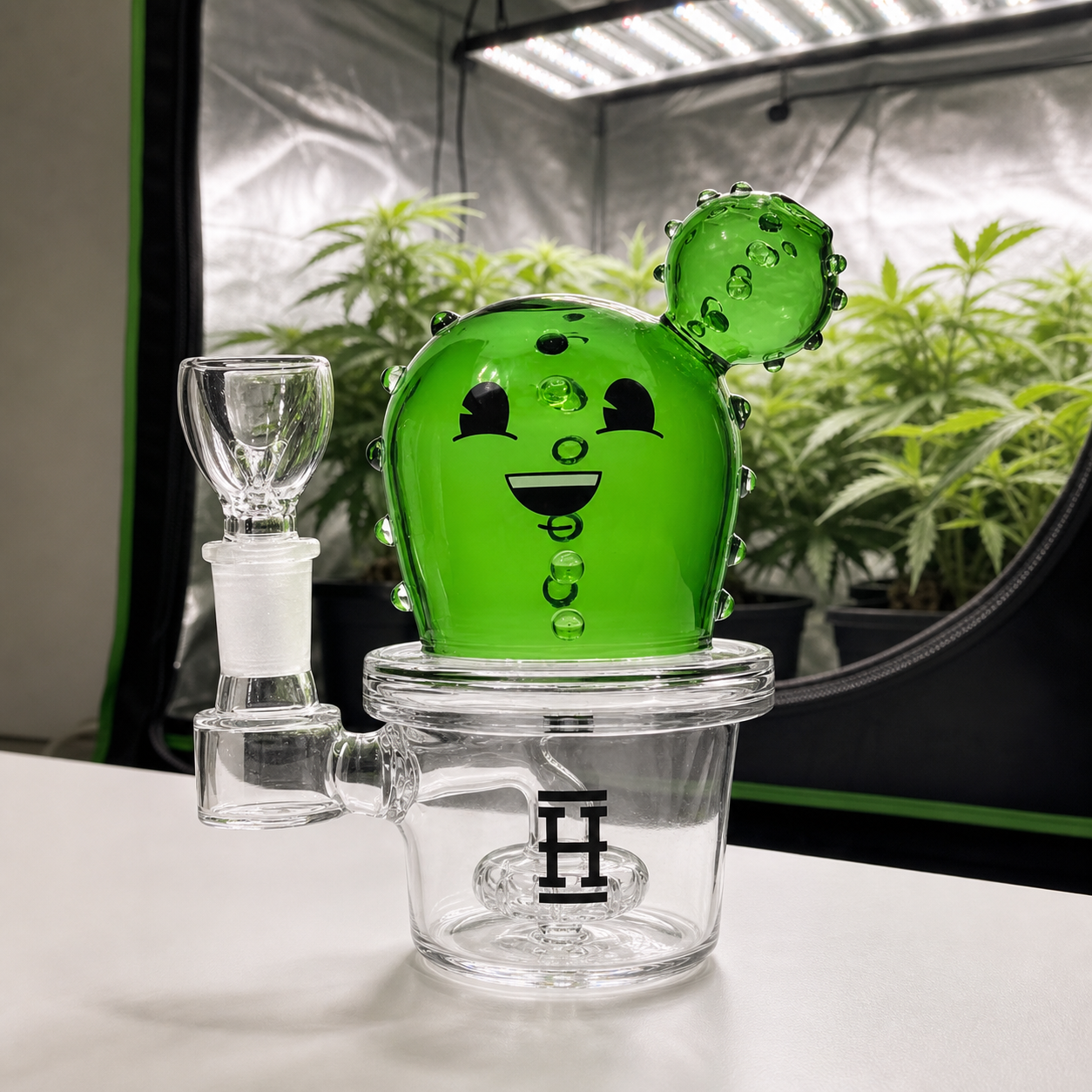 Bong Hemper Happy Cactus con cuerpo verde sonriente, base transparente y cazoleta lateral, frente a un armario con plantas.