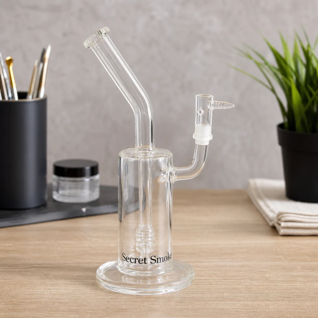 Pipa BHO Secret Smoke transparente sobre una mesa, con cámara cilíndrica, base circular, boquilla curvada y conexión lateral. Pipa BHO Secret Smoke transparente sobre una mesa, con cámara cilíndrica, base circular, boquilla curvada y conexión lateral.