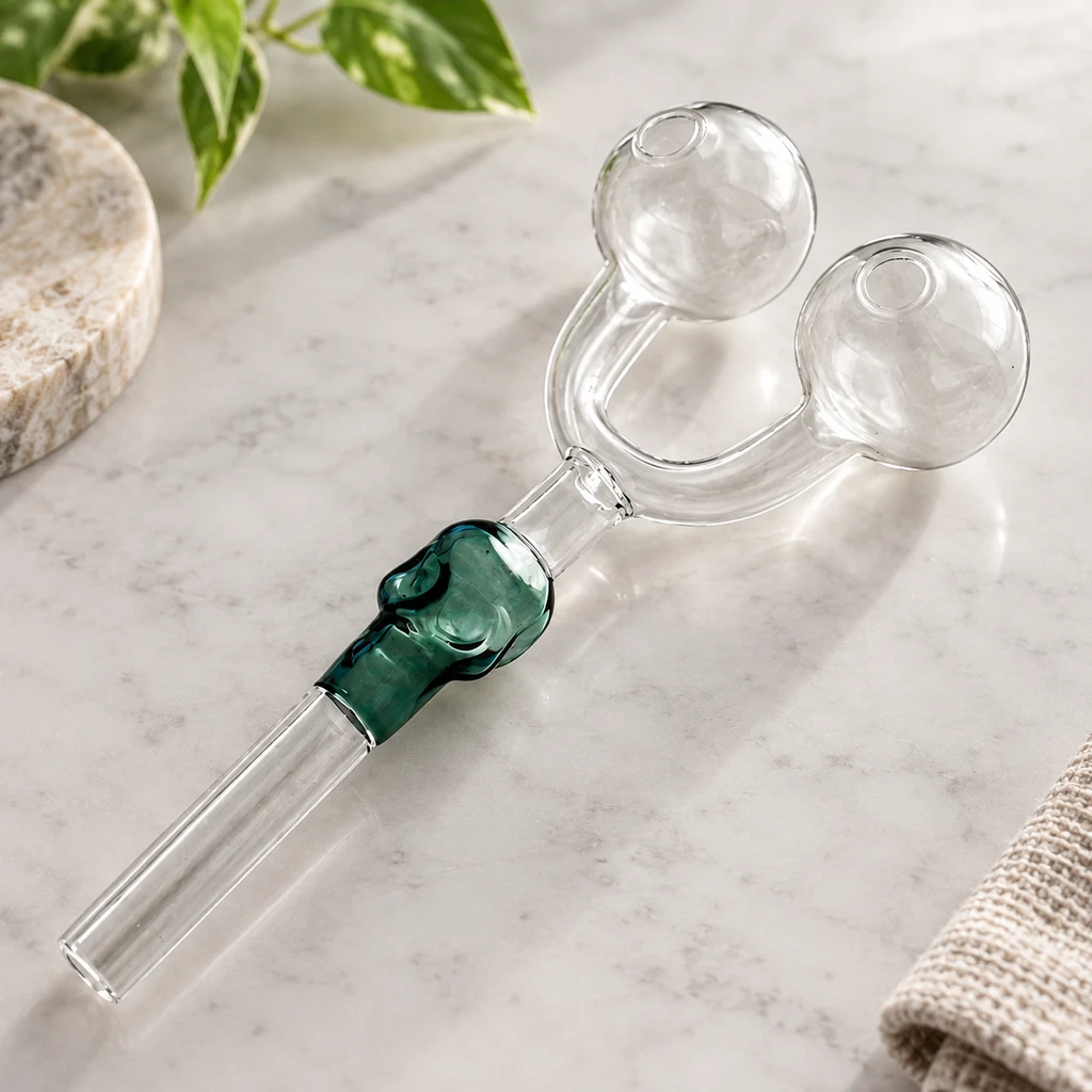 Pipa de cristal transparente con dos cámaras esféricas, tubos curvados y detalle verde junto al tubo largo, sobre una superficie clara.