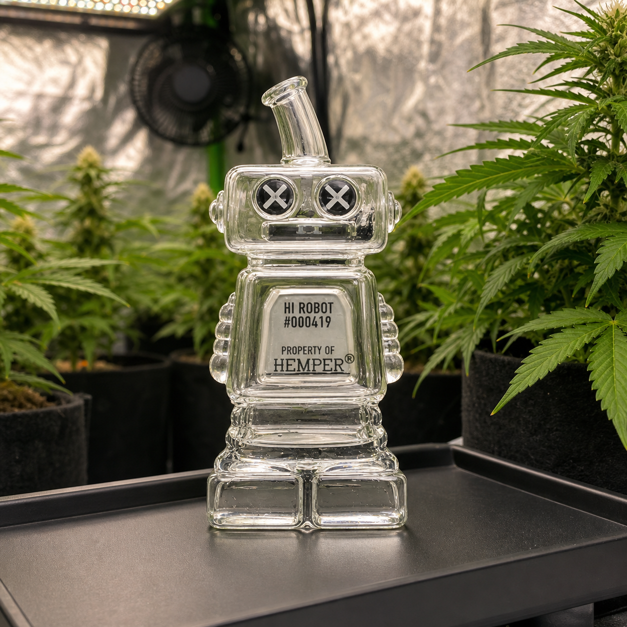 Pipa de cristal Hemper Hi-Robot con forma de robot y ojos en aspa, sobre una bandeja entre plantas de cannabis.