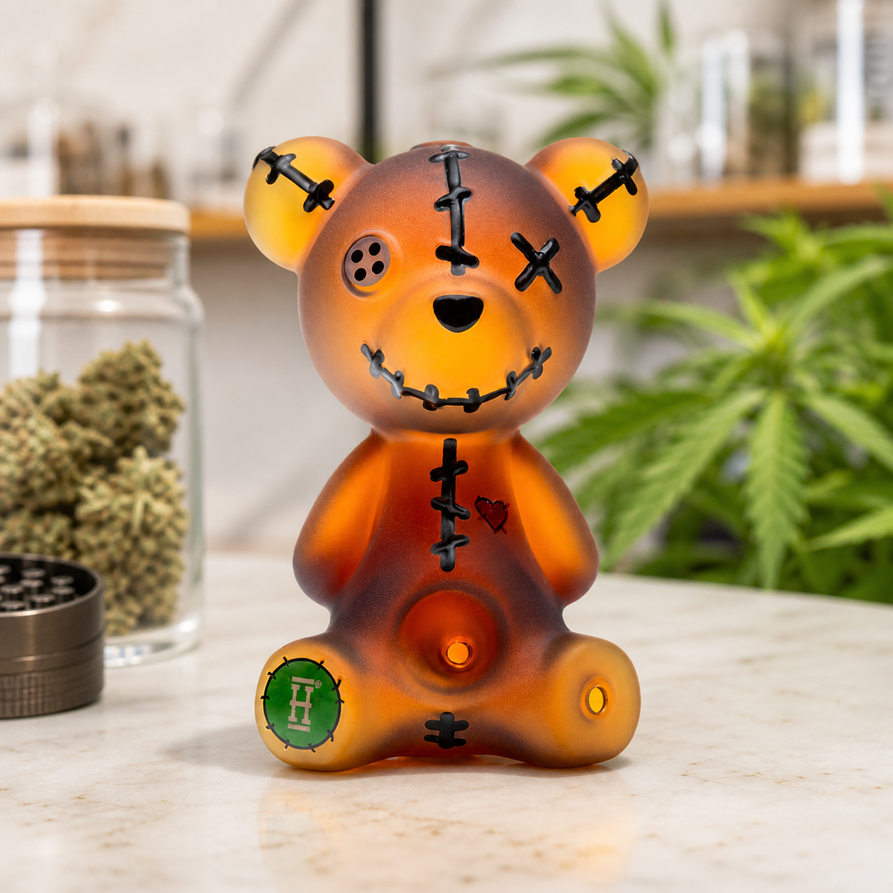Pipa Hemper Scare Bears en forma de oso naranja con costuras negras, sobre una mesa junto a un tarro con cogollos y un grinder.