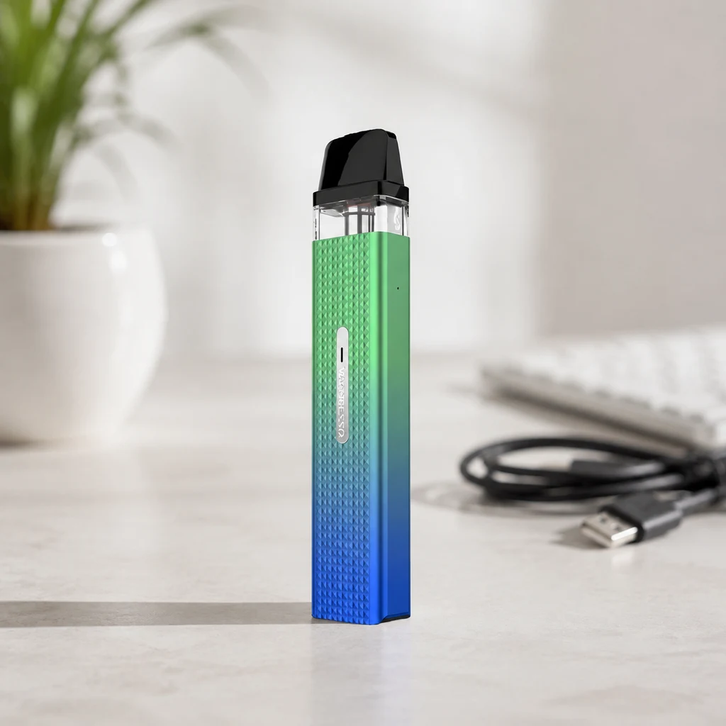 Pod Vaporesso de pie, con cuerpo texturizado en degradado verde y azul y boquilla negra; al fondo, un cable USB, un teclado y una planta.