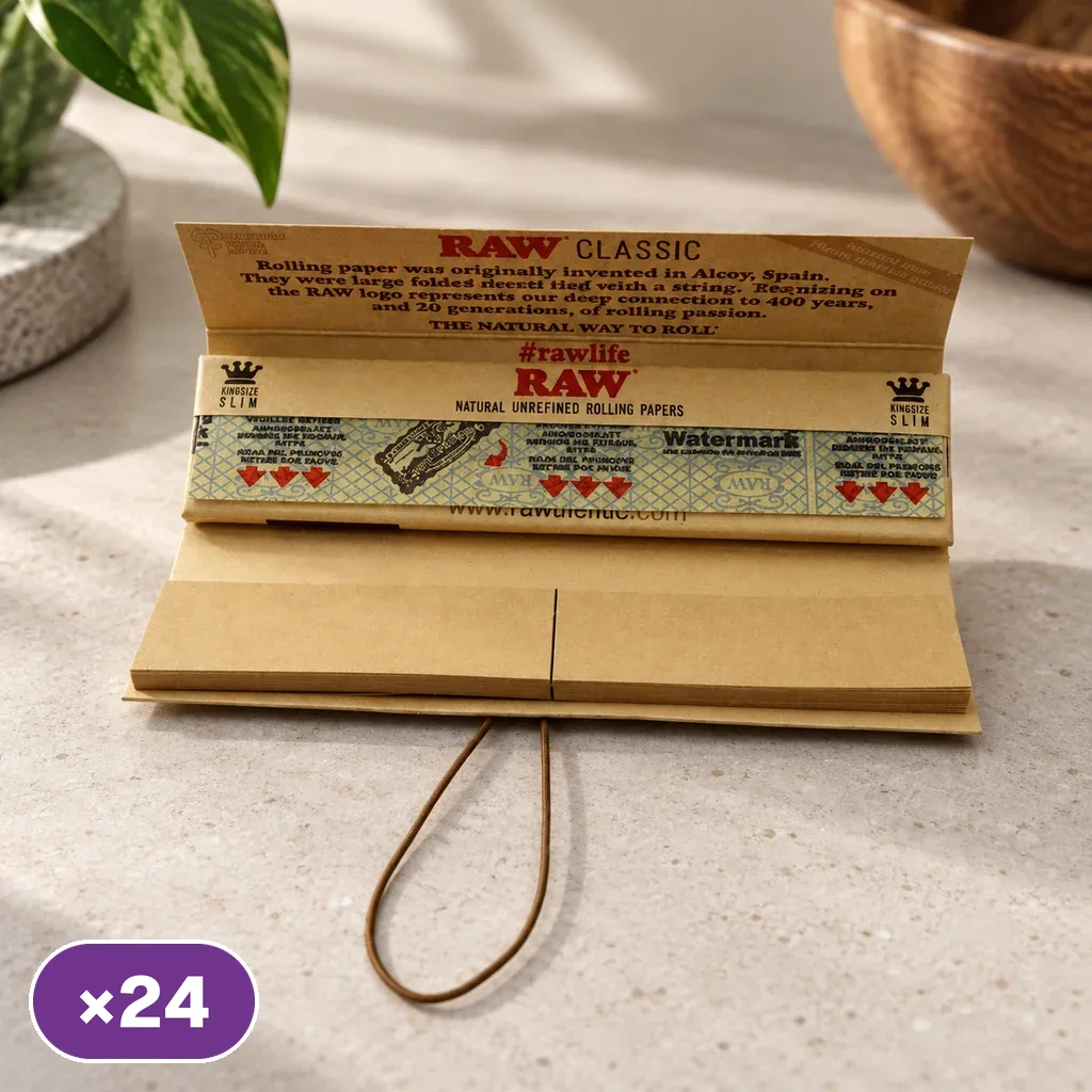 Librillo RAW abierto con papeles marrones, tips y cierre elástico sobre una mesa; la solapa muestra RAW Classic y aparece un distintivo ×24.