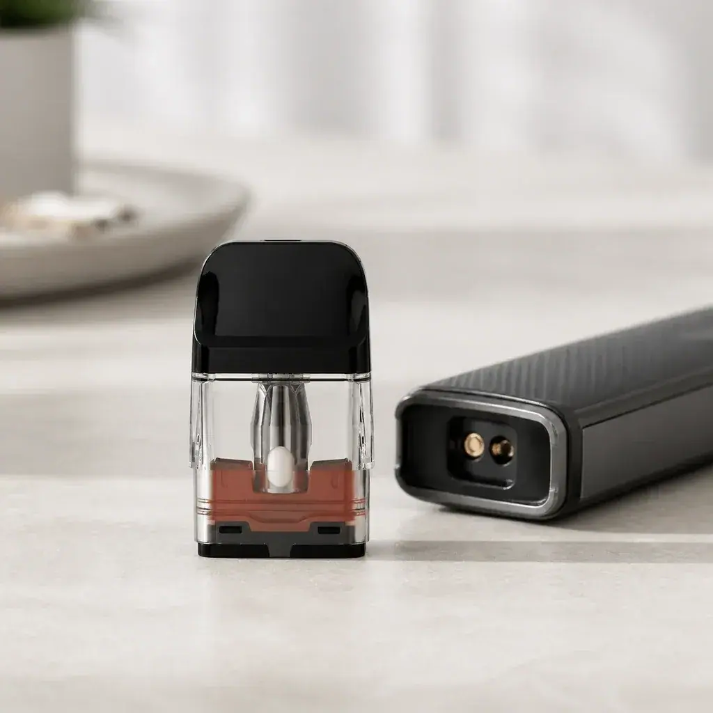 Pod recargable transparente con boquilla negra, colocado de pie junto al cuerpo de un vaporizador tumbado con los contactos visibles.