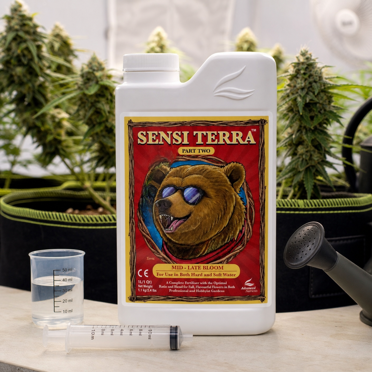 Envase de Sensi Terra Parte 2 con un oso en la etiqueta, junto a un vaso medidor y una jeringa, ante plantas de cannabis.
