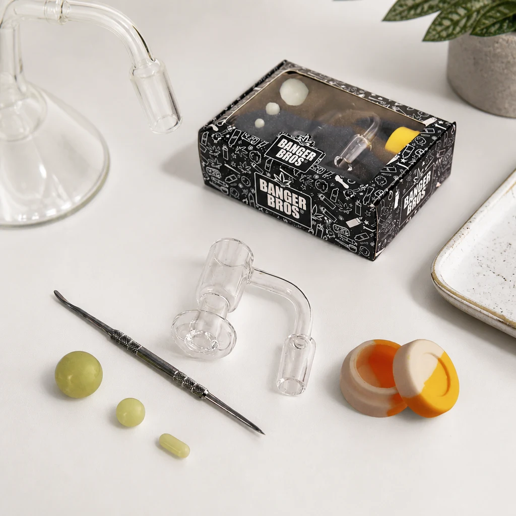 Banger transparente con tubo curvado, herramienta metálica y accesorios sobre una mesa, junto a una caja Banger Bros y un recipiente abierto.