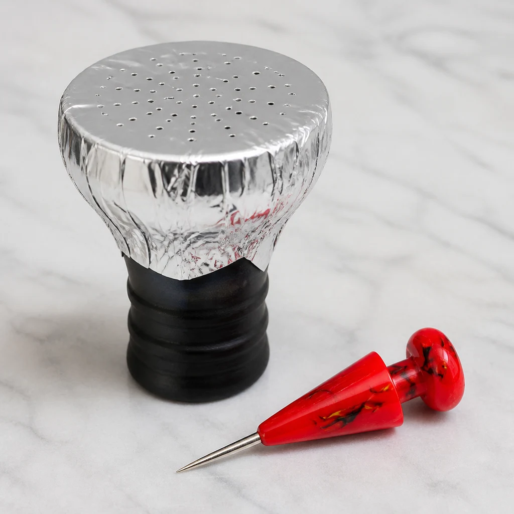 Punzón de mango rojo junto a una cazoleta de cachimba cubierta con papel de aluminio perforado, sobre una superficie clara.