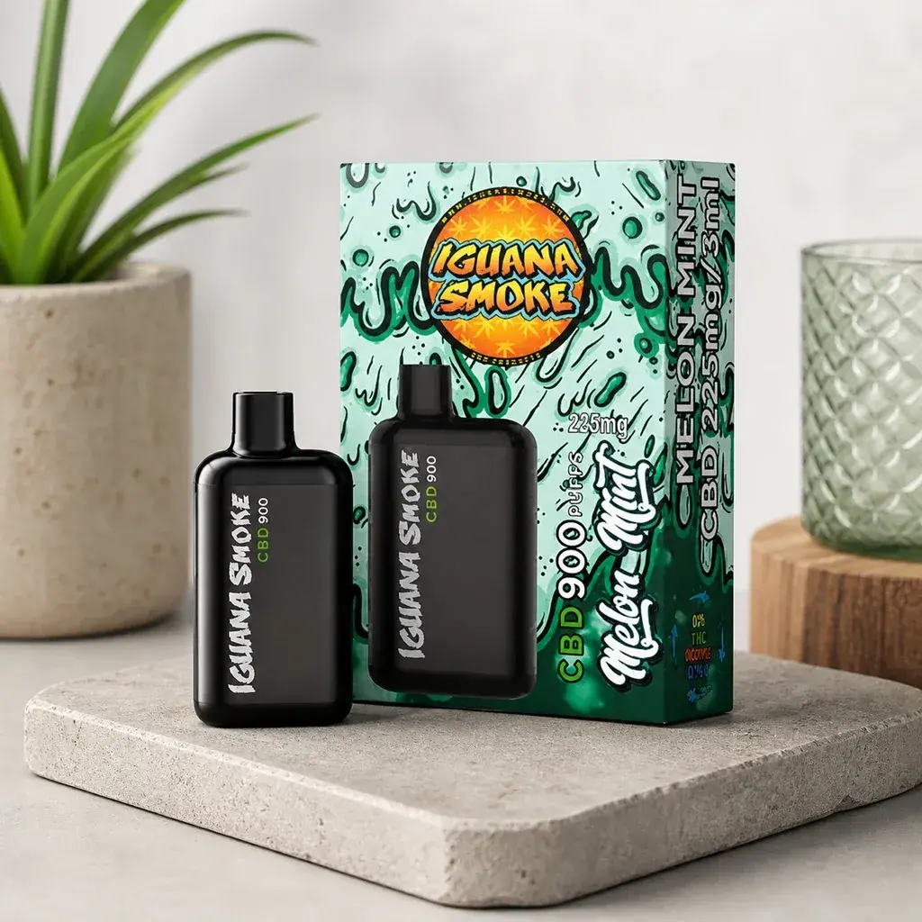 Vape negro Iguana Smoke CBD 900 Melón Menta junto a su caja verde ilustrada, sobre una base clara con una planta y un vaso al fondo.