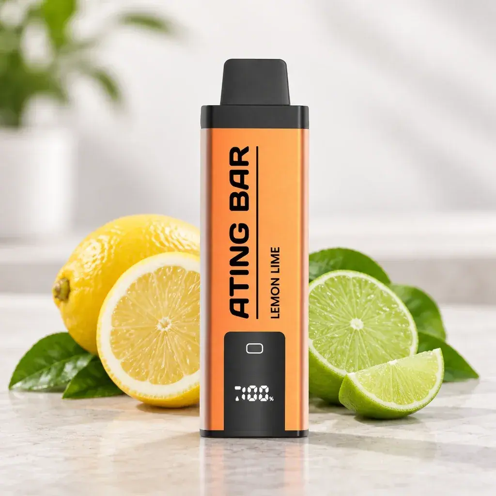 Vapeador Ating naranja con boquilla negra, pantalla e inscripción Lemon Lime, colocado entre limones y limas cortados.