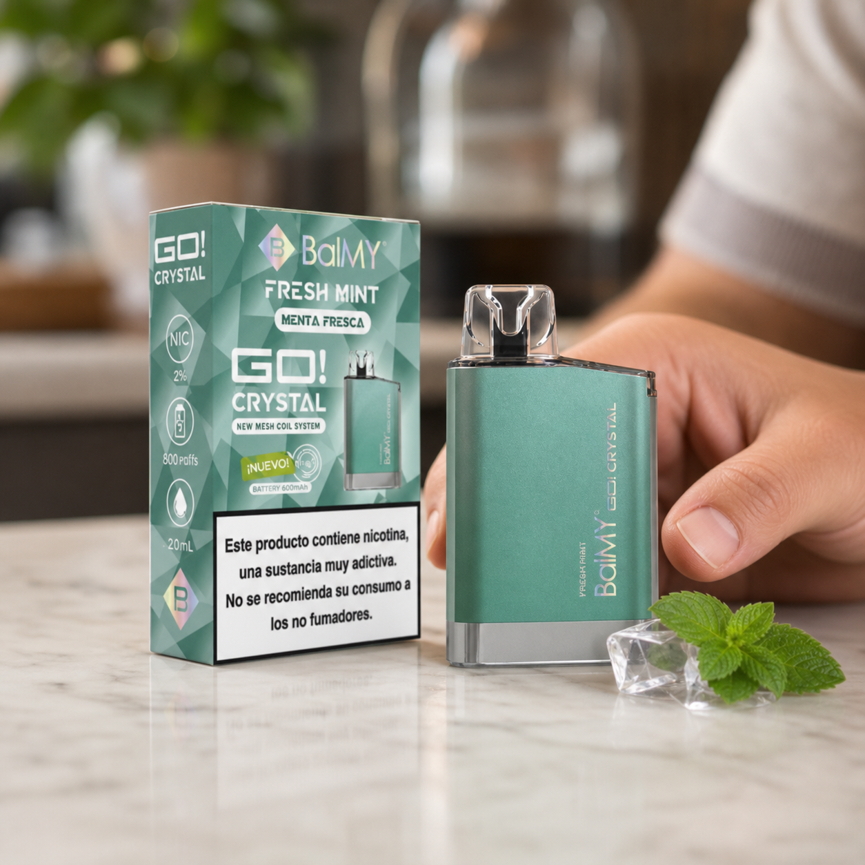 Vaper Balmy Go Crystal Fresh Mint verde junto a su caja, con una mano detrás y hojas de menta y cubos transparentes sobre la mesa.