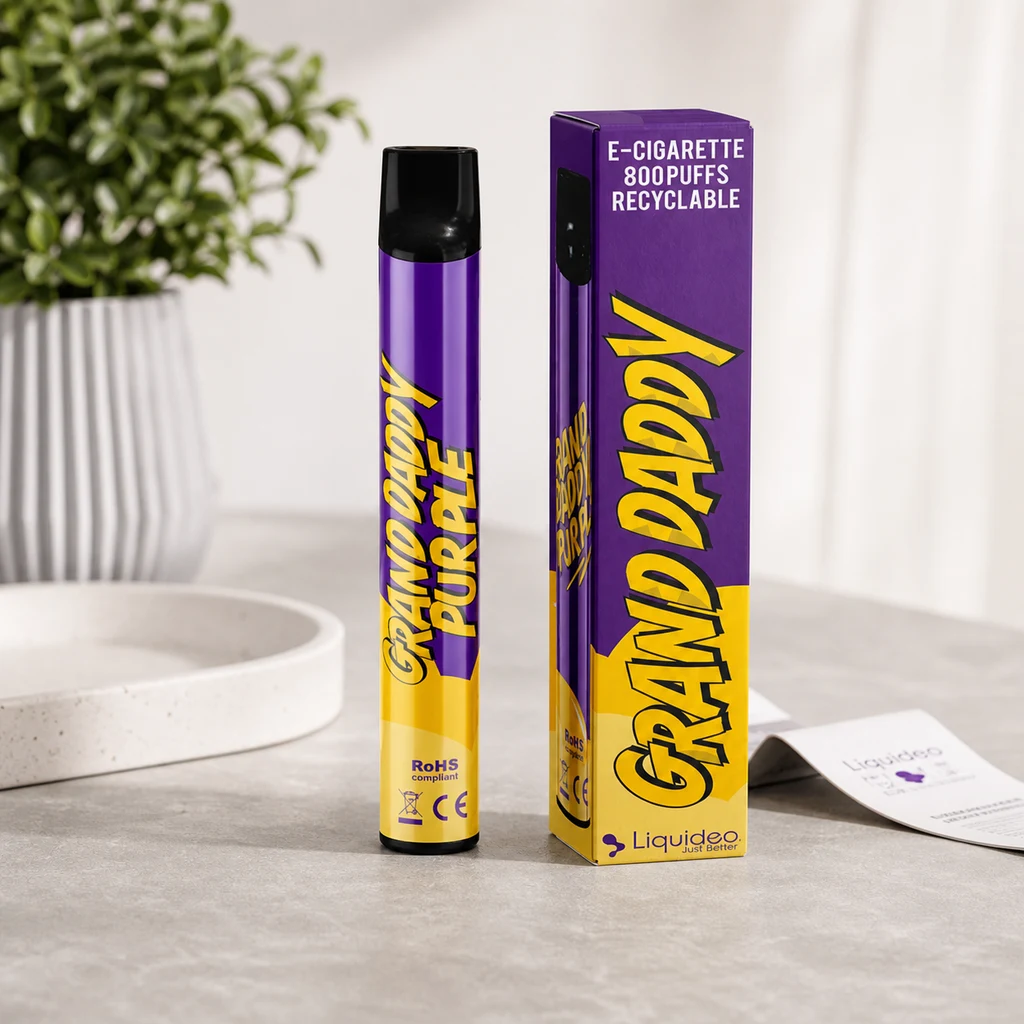 Vaporizador Grand Daddy Purple morado y amarillo junto a su caja, sobre una mesa clara con una planta al fondo.