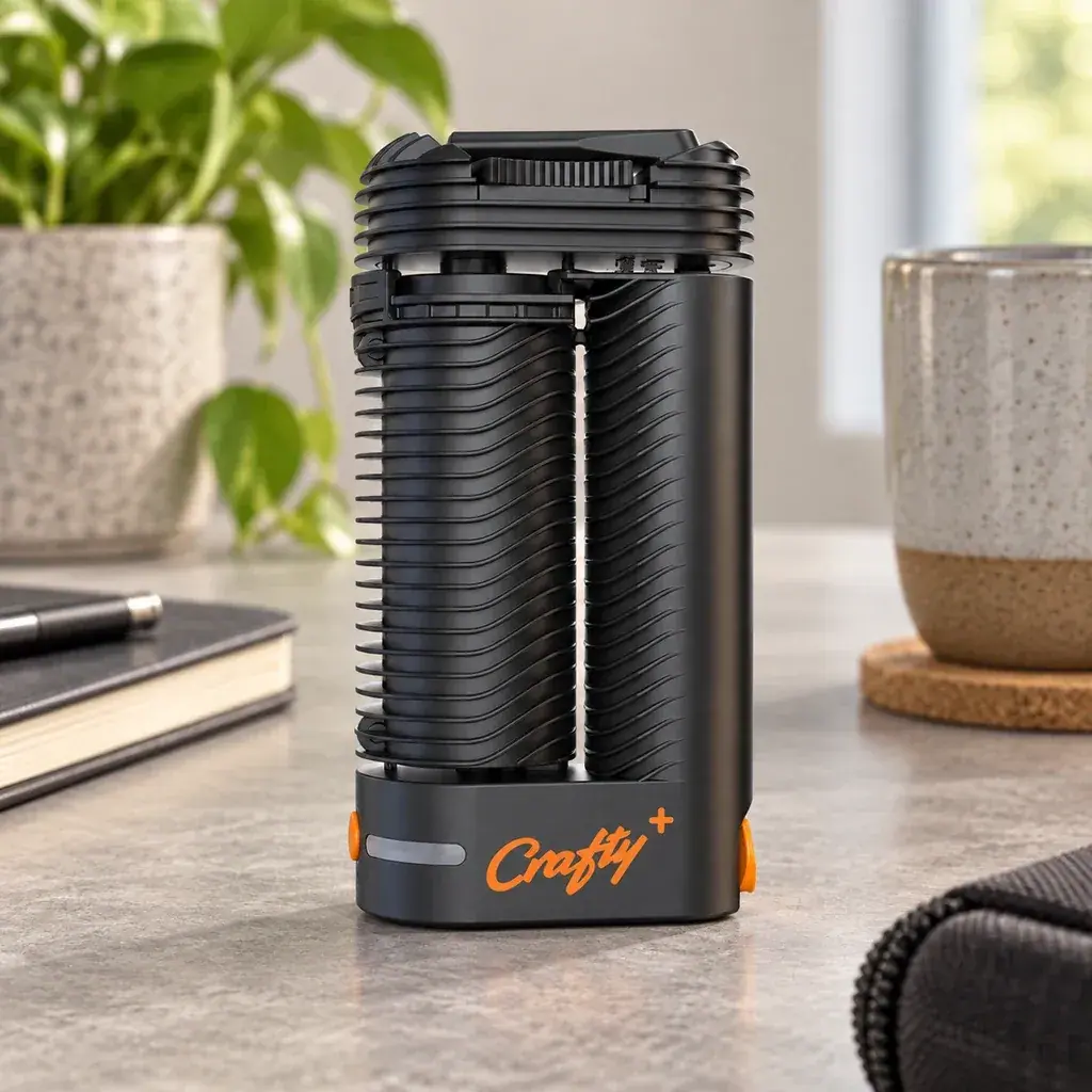 Vaporizador Crafty+ negro con cuerpo acanalado y detalles naranjas, sobre una mesa junto a una planta, un cuaderno y una taza.