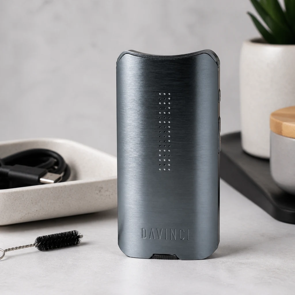 Vaporizador Davinci IQ3 Gunmetal gris oscuro en posición vertical, con puntos luminosos frontales, junto a un cepillo y un cable.