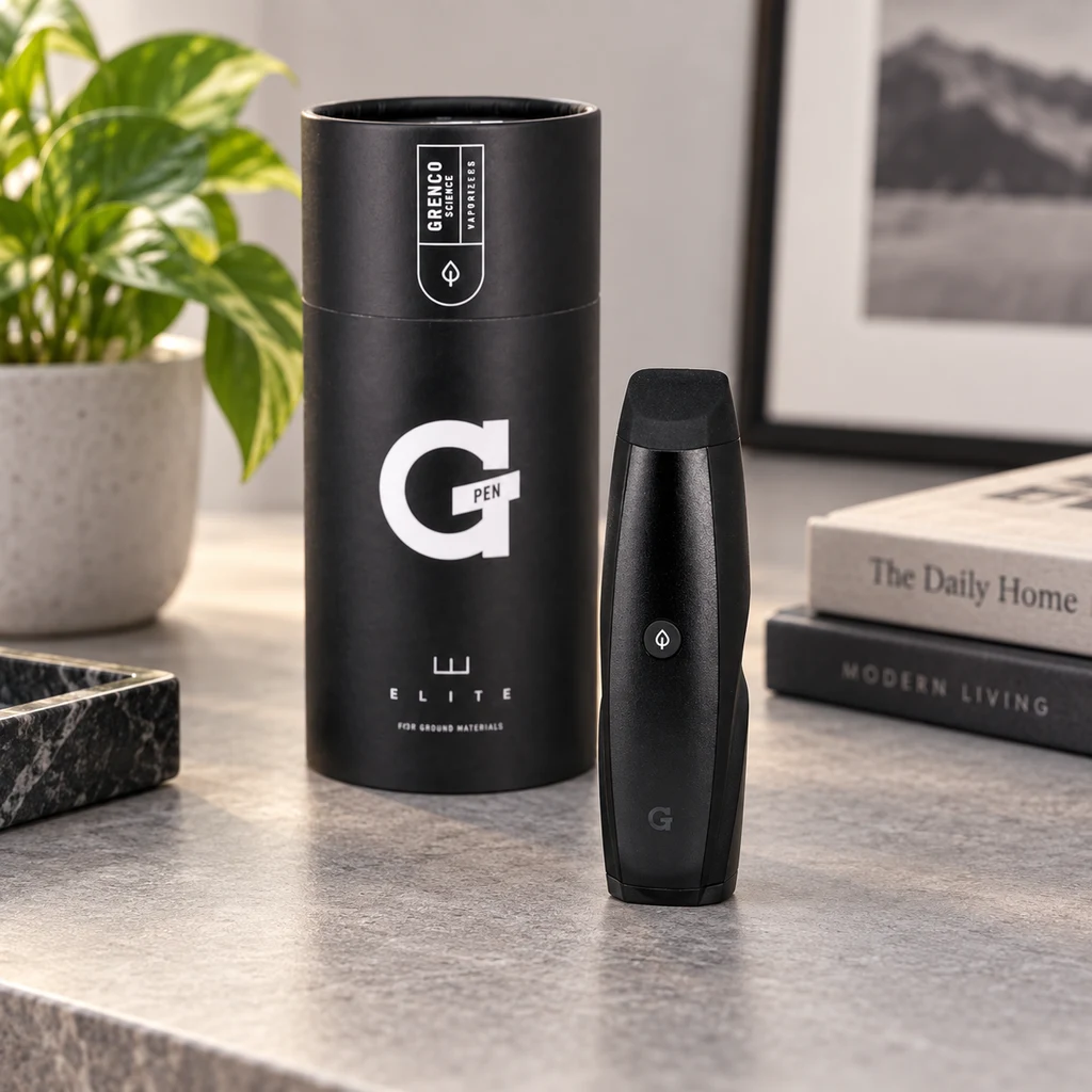Vaporizador negro G-Pen junto a su envase cilíndrico sobre una mesa, con una planta y libros al fondo.