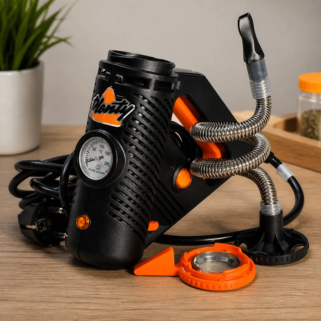 Vaporizador PLENTY negro y naranja con termómetro frontal, tubo metálico enrollado y boquilla, con accesorios sobre una mesa de madera.