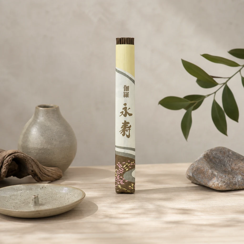 Tubo de incienso Kyara Eiju con caracteres japoneses y bastones asomando, sobre una mesa junto a un incensario, un jarrón y una piedra.