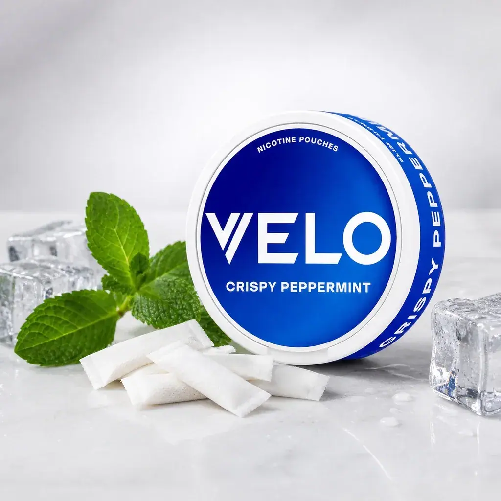 Envase redondo azul y blanco de VELO Crispy Peppermint junto a bolsas blancas, hojas de menta y cubitos de hielo.