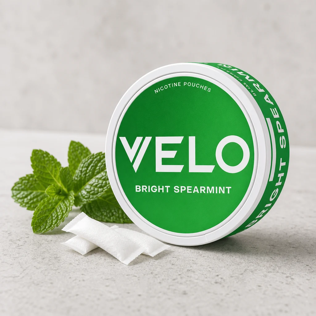 Envase redondo blanco y verde de VELO con texto Bright Spearmint, junto a bolsitas blancas y hojas de menta. Envase redondo blanco y verde de VELO con texto Bright Spearmint, junto a bolsitas blancas y hojas de menta.