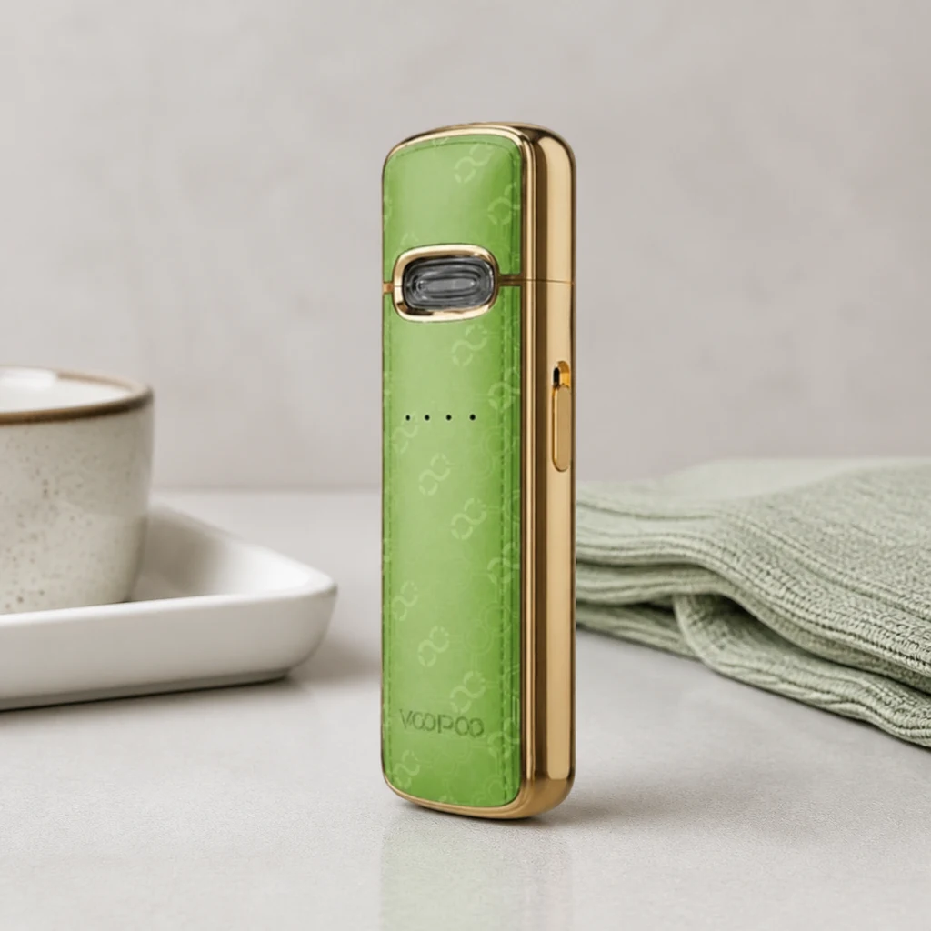 Voopoo Vmate E verde con bordes dorados, botón frontal e indicadores, colocado de pie junto a una taza y una tela verde.